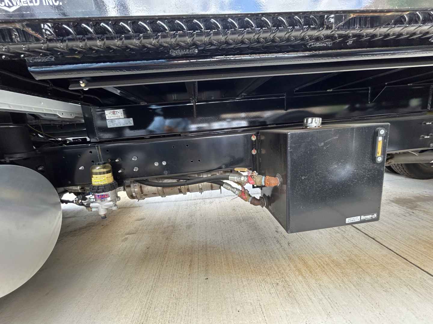 Underbody view of a 2026 Mack MD6 flatbed truck showing a black rectangular toolbox mounted to the frame with hoses and fittings nearby and a yellow cap on a reservoir.