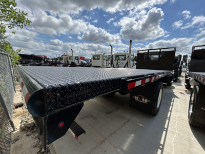 Flatbed trailer chassis of a 2026 Mack MD6 truck showing diamond plate deck and wheel area with spare tires in background.