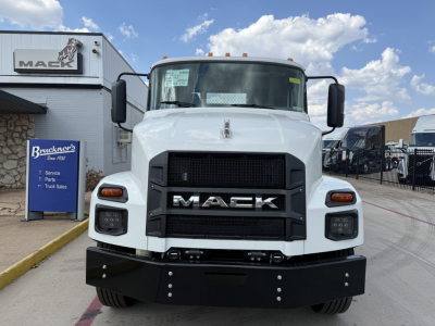 Front view of a white 2026 Mack MD6 truck showing bold black grille with MACK lettering, large black bumper, side mirrors, and factory sticker on windshield.