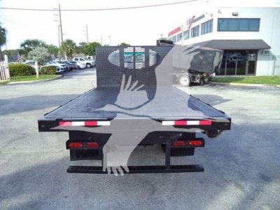 Isuzu NPR flatbed truck rear view showing flatbed platform and taillights, yard with building in background.