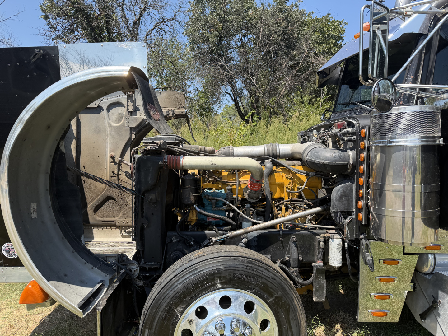 Peterbilt 379X engine bay with exposed diesel engine, intake pipes, hoses, and front wheel visible; side view of cab and chrome air cleaners.