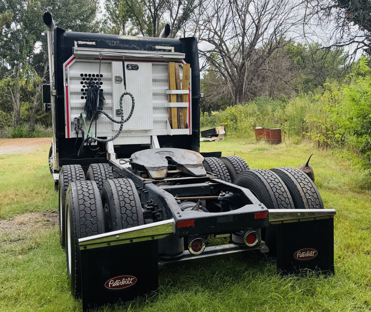 Peterbilt 379X conventional sleeper truck chassis rear view showing fifth wheel, dual rear axles, mud flaps with Peterbilt logo, and power unit area in a grassy lot.