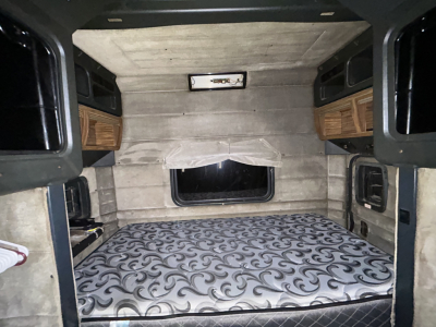 Peterbilt truck cab interior showing sleeping area with patterned mattress, overhead cabinets, side walls, and window; no visible branding or part numbers in the image.