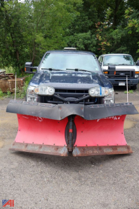 Ford F-350 Super Duty pickup with a large front plow attached, blue exterior, two headlights visible, truck in a parking lot with trees in background.