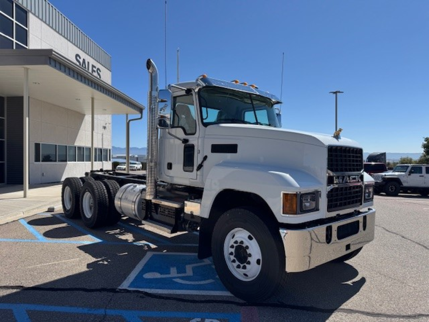White Mack PI64 truck in a dealership parking lot, cab with sleeper, dual rear axles, front grille, and side exhaust. No visible text on the truck itself.