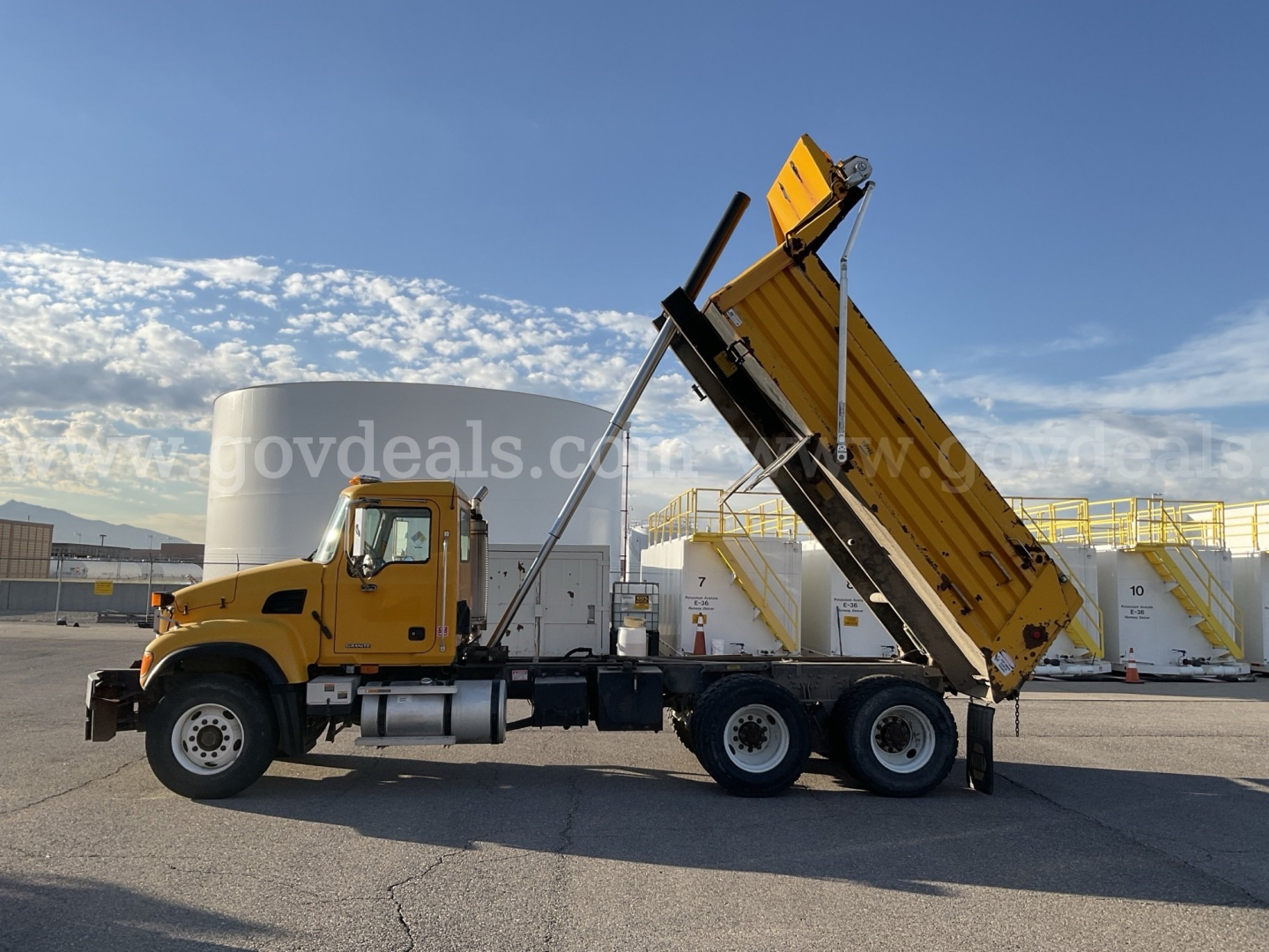 2005 Mack CV713 dump truck in service position with dump bed raised, yellow cab and chassis, side view, with silos in background.