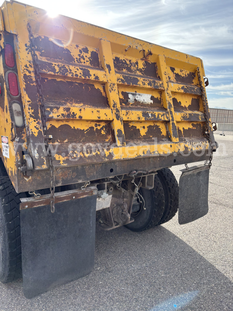 Back of a yellow dump truck bed with heavy wear showing rust and paint chipping, Mack CV713, dual rear tires, mud flaps, hydraulic components visible.
