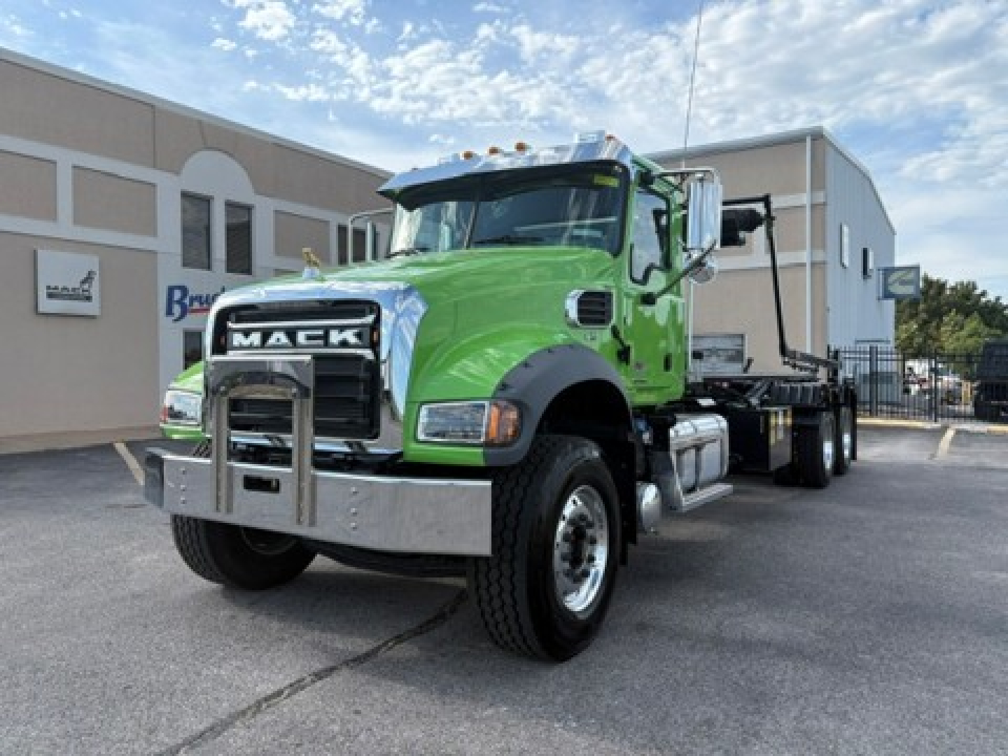 Green 2026 Mack GR64F rolling truck with chrome bumper and flatbed in a dealership lot. Visible Mack grille, large tires, cab with side mirrors, and emission exhaust behind.