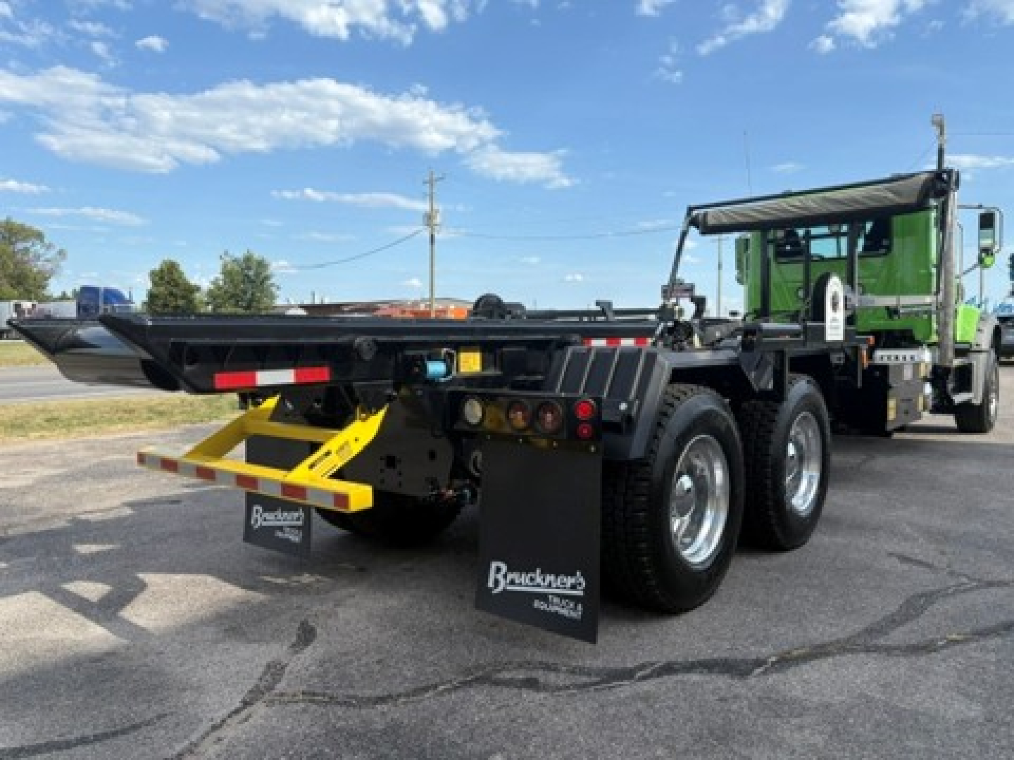 Rolloff truck rear view showing black dump body with yellow ramps and mud flaps reading Bracken's Roll-Offs; green cab in background under clear sky.