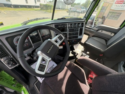Mack GR64F cab interior showing steering wheel, dashboard gauges, and control panel with gear selector; green truck exterior visible at edge.