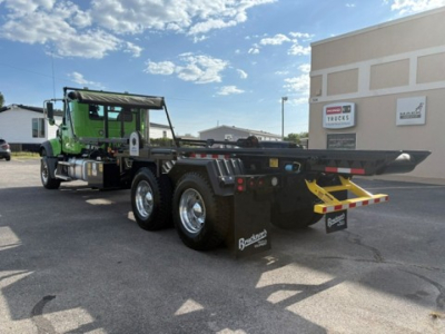 Green Mack GR64F roll-off truck in a lot, rear view showing dual rear axles, heavy-duty flatbed, towing/yank bar, and mud flaps with branding.
