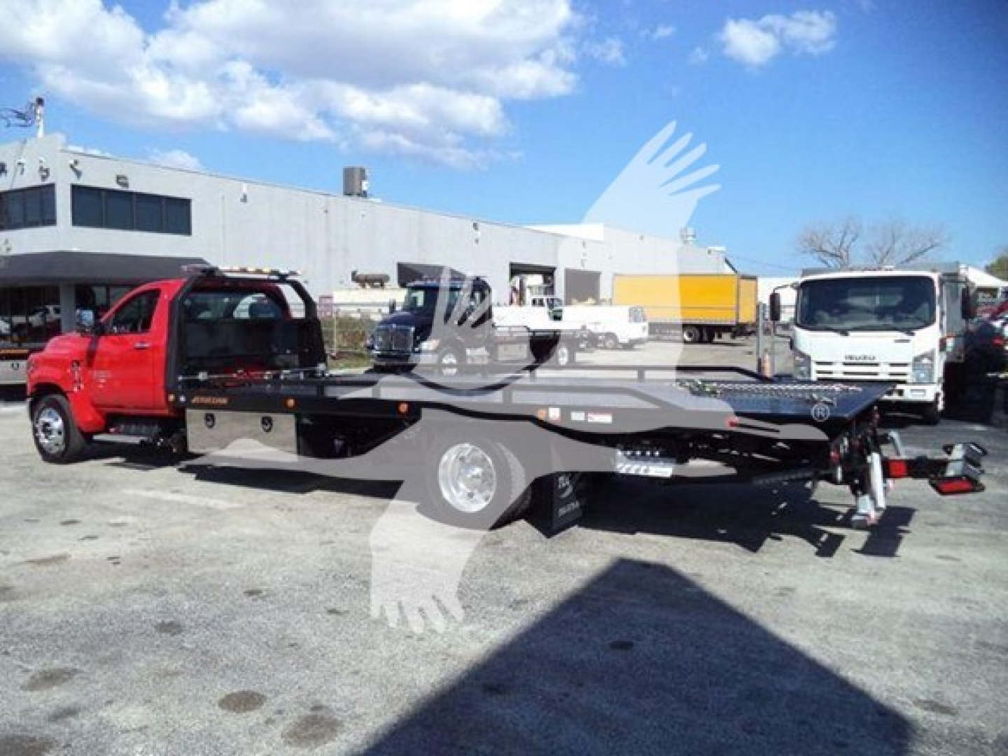 Flatbed rollback tow truck with a red cab and black flatbed, parked outdoors in a lot with a white industrial building in the background.