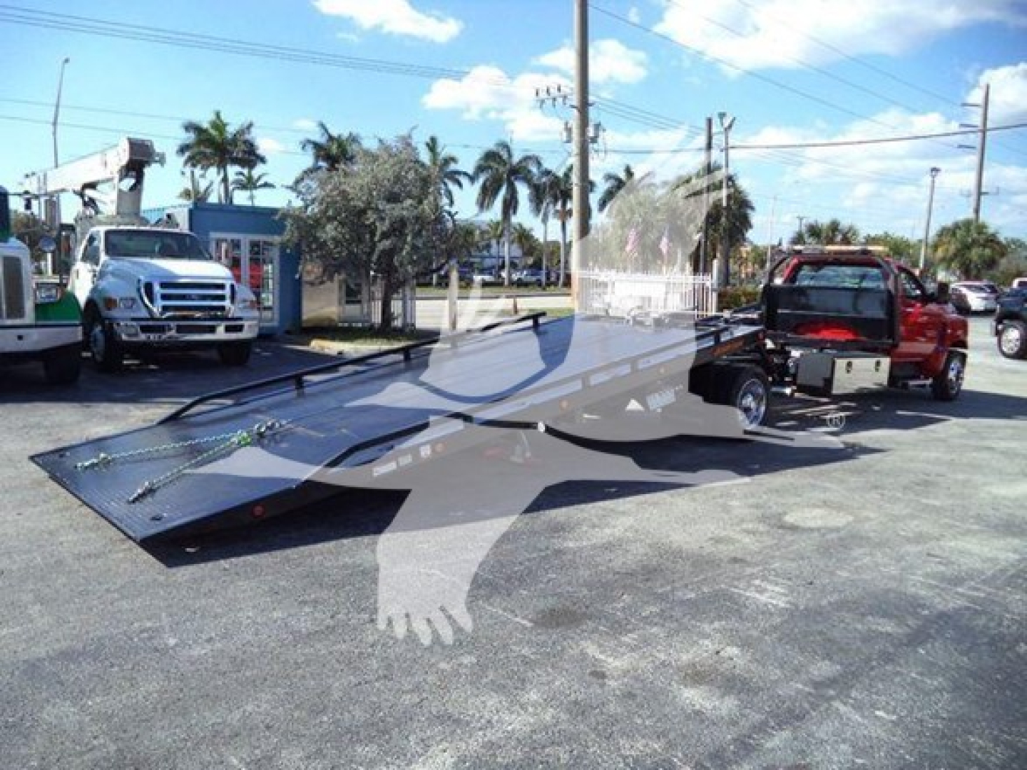 Truck bed of a rollback tow truck attached to a red tow vehicle, in a dealership lot with other trucks and palm trees in the background.