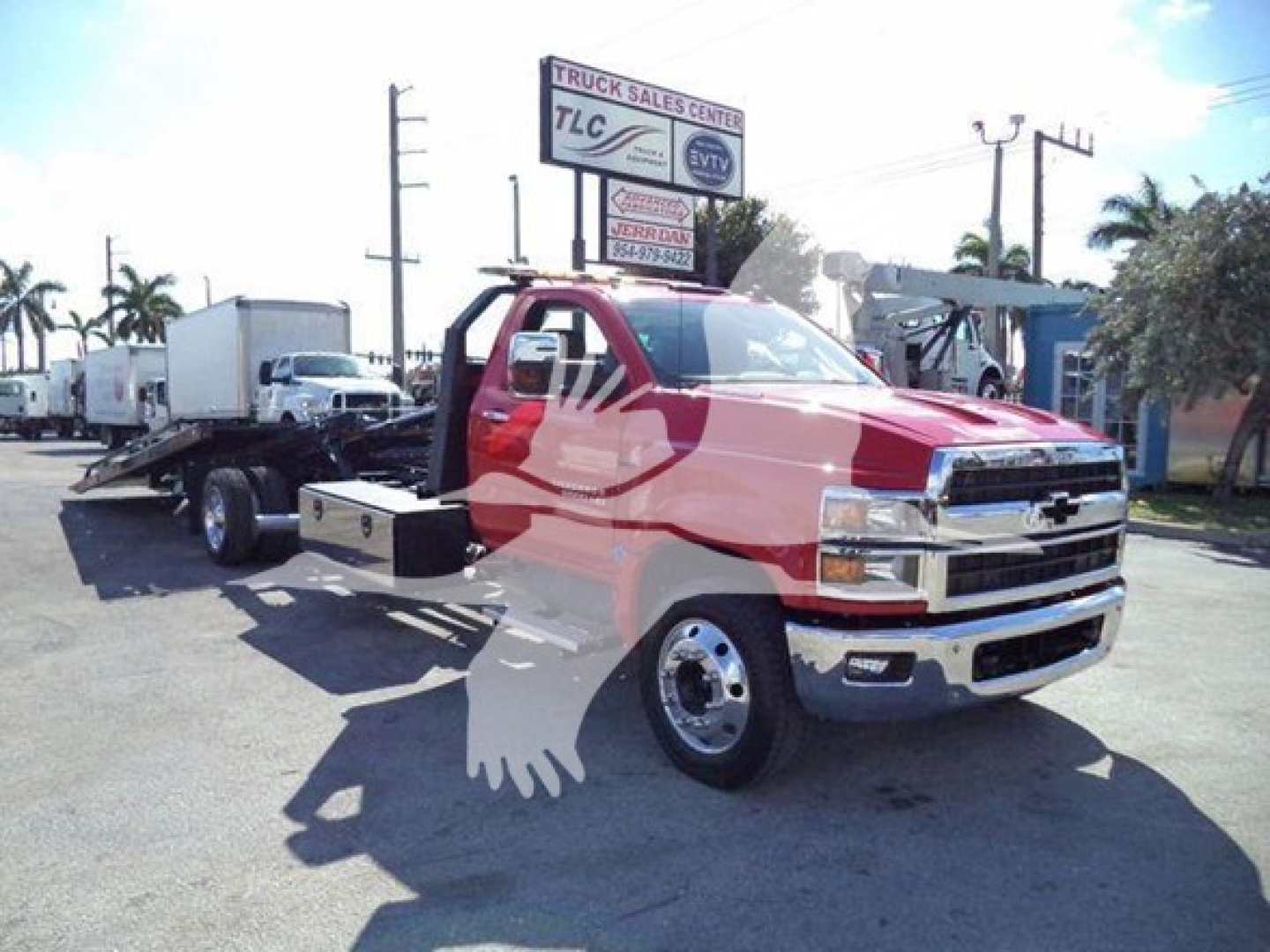 Truck tow rig in a dealership lot showing a red and white 2023 Chevrolet Silverado 6500HD tow truck with a long wheelbase and flatbed, dealership signs visible in background.