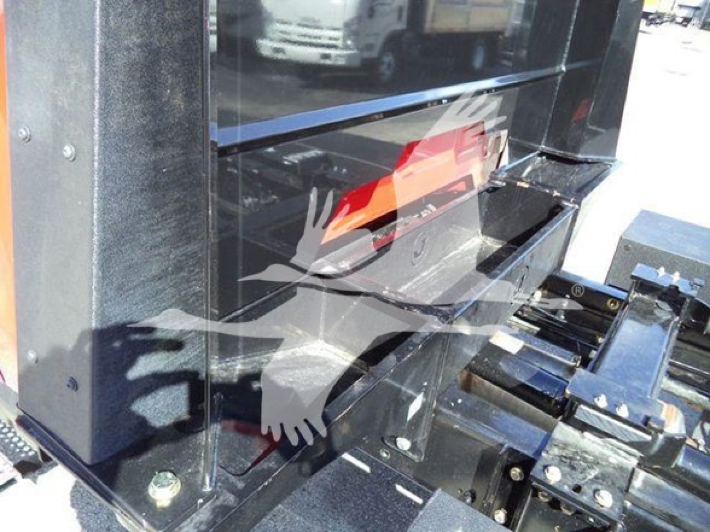 Truck rollback equipment: black metal toolbox/compartment with chrome arms and hydraulic mechanism, reflective surface showing surrounding vehicles.