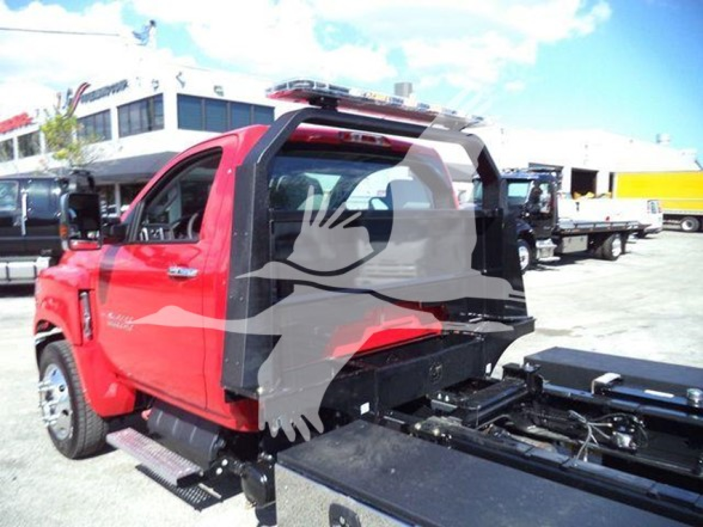 Truck cab and rollback chassis of a 2023 Chevrolet Silverado 6500HD seen from rear-left, red body, with flatbed behind cab and dealership lot in background.