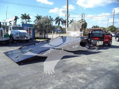 Truck bed of a rollback tow truck attached to a red tow vehicle, in a dealership lot with other trucks and palm trees in the background.