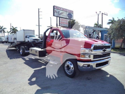 Truck tow rig in a dealership lot showing a red and white 2023 Chevrolet Silverado 6500HD tow truck with a long wheelbase and flatbed, dealership signs visible in background.