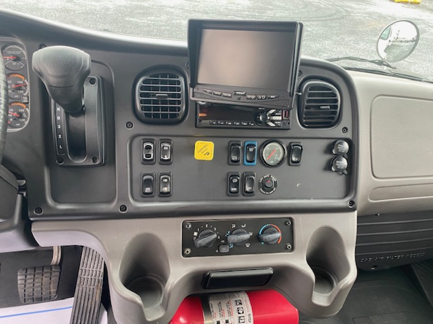 Cab interior of a 2019 Freightliner M2-106 showing steering column, gear selector, control switches, mounted monitor, and HVAC dials.