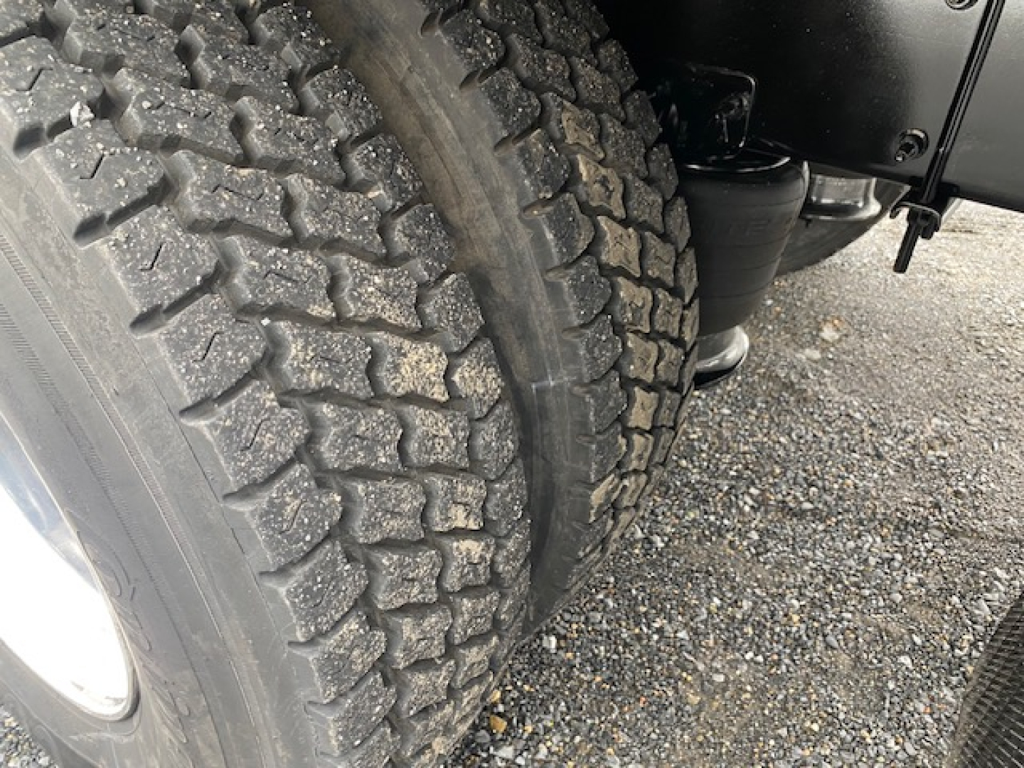 Truck wheel and dual tires on Freightliner M2-106, heavy-duty tire tread pattern visible; part of front axle and undercarriage with gravel surface.