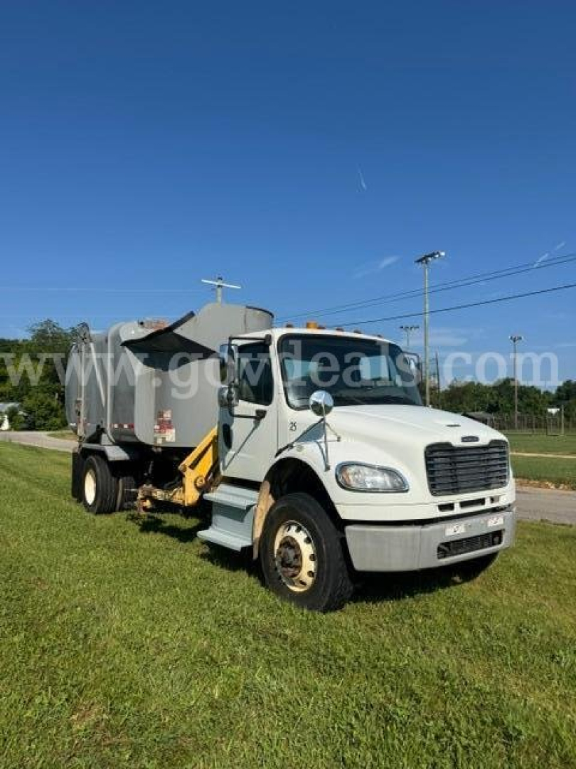 Truck in service bay showing Freightliner M2 with garbage body and side grabber on rear, white cab, front view.