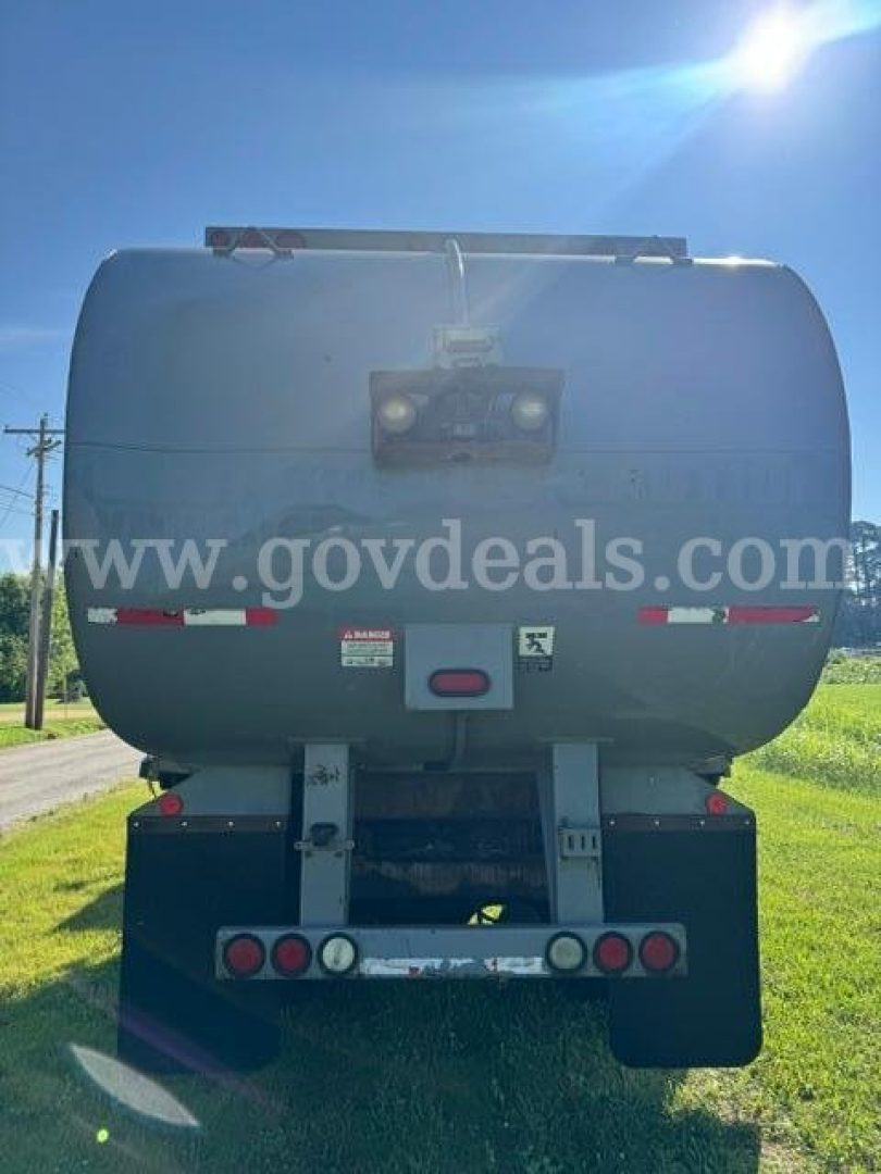 Rear view of a Freightliner M2 dump/garbage truck showing a gray tank, taillights, bumper, and frame; visible stickers on the tank and a reflective strip.