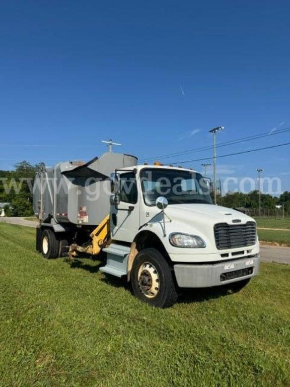 Truck in service bay showing Freightliner M2 with garbage body and side grabber on rear, white cab, front view.