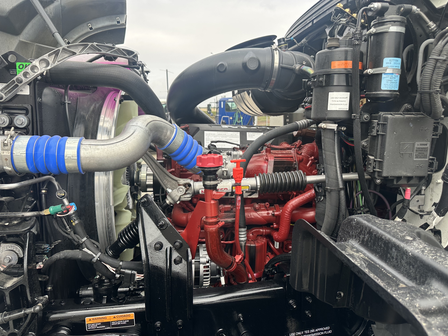 Mack GR64F engine bay with red engine block, blue hose, and various black components; visible fuel/air lines and coolant hoses; compact layout of truck engine compartment.