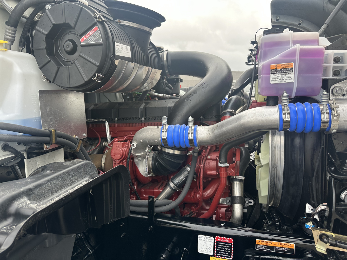 Mack GR64F engine bay showing diesel engine with red block, air intake, blue silicone hoses, coolant reservoir, and auxiliary components.