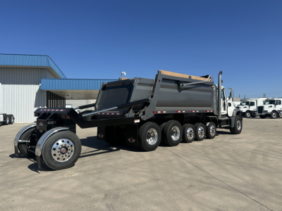 Mack GR64F dump truck in a lot, side view showing rear dump bed and multiple axles with six wheels, white cab, metallic gray bed, blue sky.
