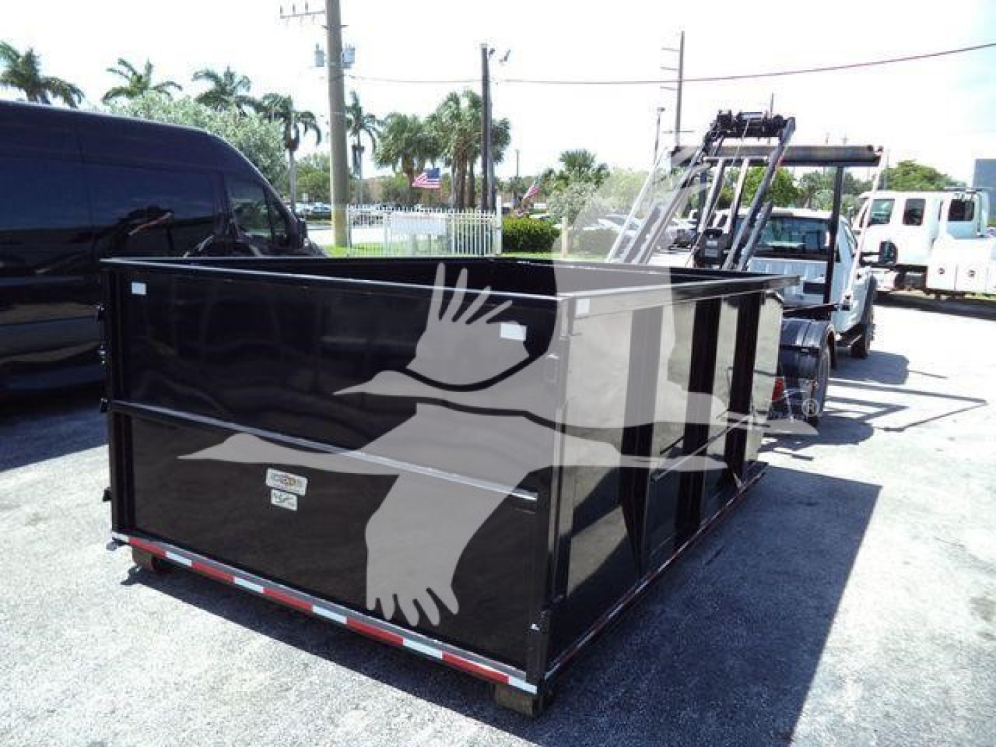 Flatbed roll-off truck bed with black metal sides and white bird graphic, attached to a tow vehicle in a lot.
