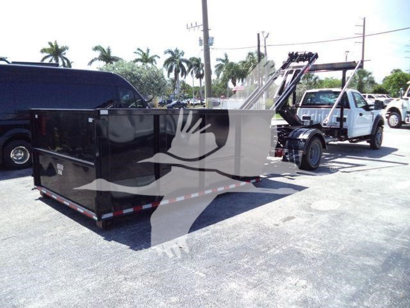 Truck-mounted roll-off body in a parking lot with a crane arm and a black rectangular dumpster container attached. Ford F-550 likely used, with other trucks in background.