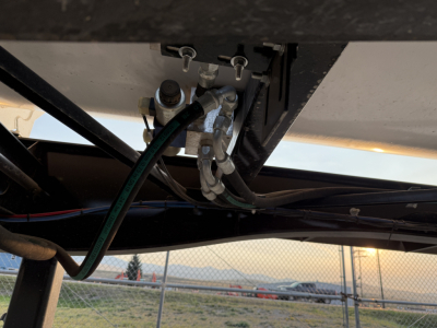 Trailer undercarriage showing a dual hose quick-connect coupling and wiring harnesses; visible metal brackets and a light blue painted surface behind.