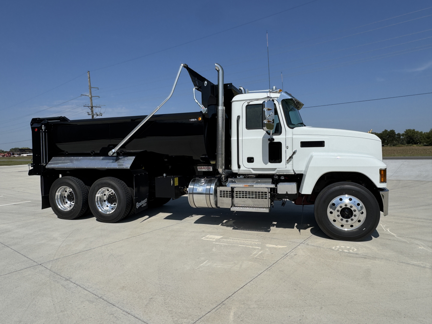Truck: Mack PI64 dump truck side view showing black dump bed, white cab, dual rear axle, chrome fuel tank and steps.