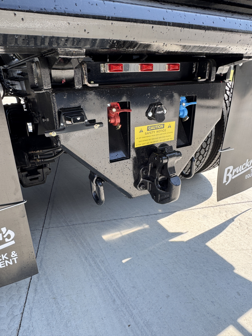 Rear chassis and hitch area of a new Mack PI64 dump truck showing control levers, warning label, and electrical connectors.