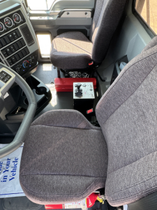 Cab interior of a new 2026 Mack PI64 dump truck showing configured driver's seat and center console with a red battery or control module in the floor area.