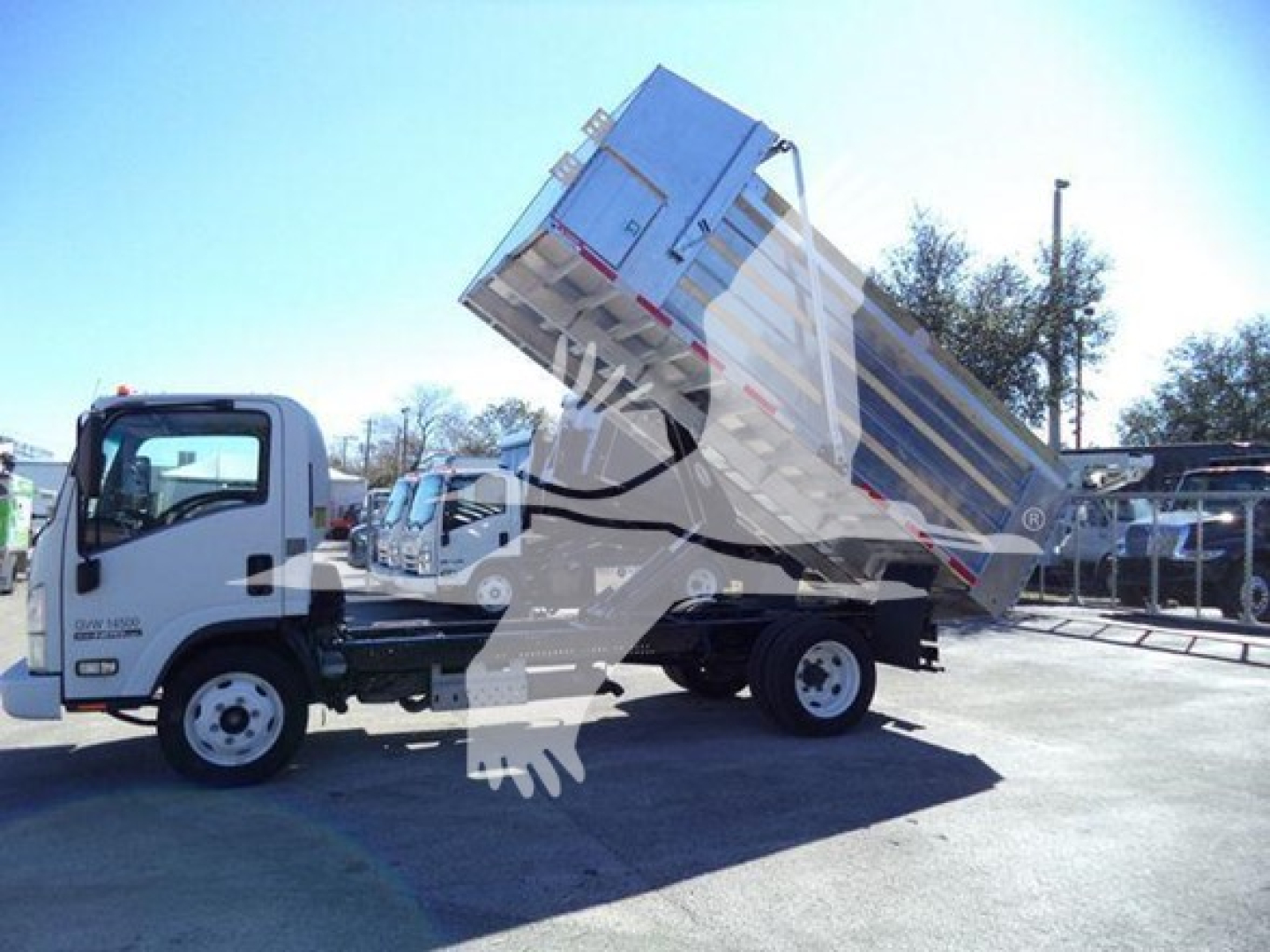 Isuzu NPR-HD dump truck in operation on a lot, side view with bed raised, displaying truck cab and dumping cargo bed.