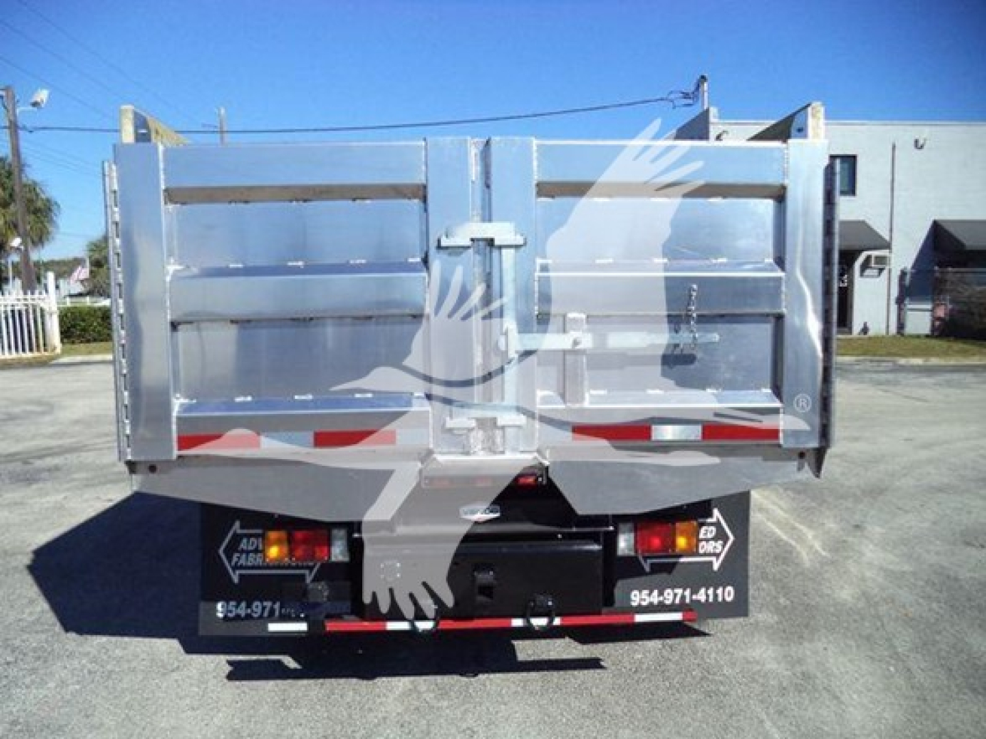 Isuzu NPR HD dump truck rear view showing metal cargo bed with tailgate and rear lights. Visible text includes phone numbers and dealership branding on bumper.