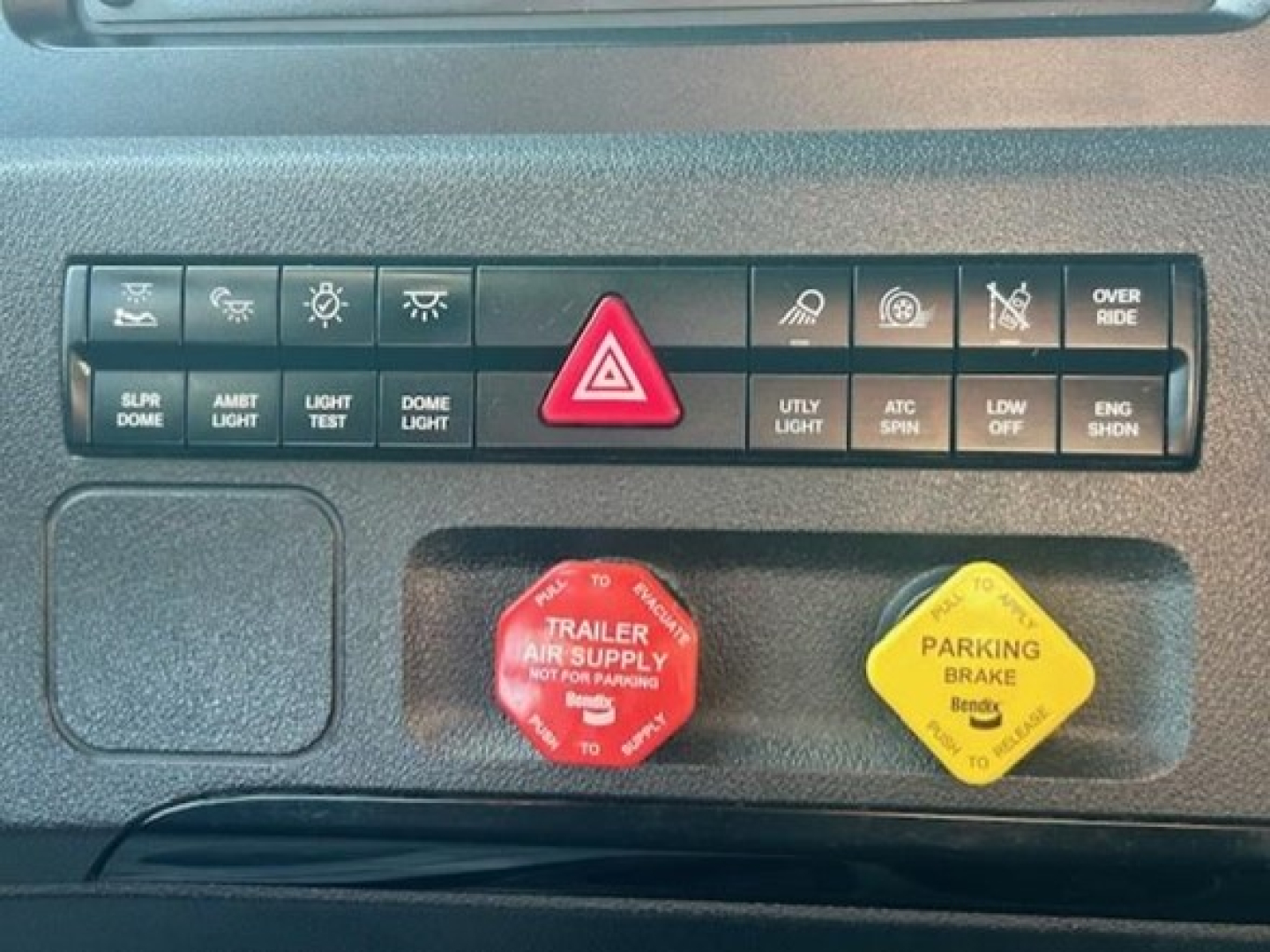 Dashboard switches and warning indicators in a Freightliner Cascadia cab. Visible items include hazard triangle, left and right toggle buttons, red trailer air supply knob, yellow parking brake knob, and various icons labeled on the control