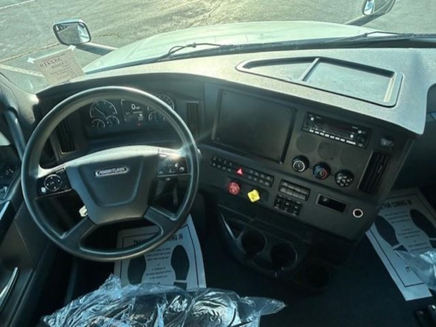 Cab interior of a Freightliner Cascadia sleeper with steering wheel, dashboard, and controls visible; driver seat area and paperwork on the floor.