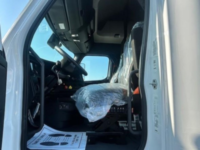 Cab interior of a 2023 Freightliner Cascadia 126 tractor viewed from open driver door; plastic-wrapped driver's seat; steering wheel and dash visible; light gray exterior; panoramic view of cab interior.