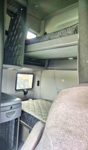 Truck sleeper cab interior showing upper bunk with mattress and lower bunk, small window, and storage cabinet.