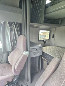 Truck cab interior showing sleeper layout with bunk bed area, storage compartments, and a small window; driver seat visible in foreground.