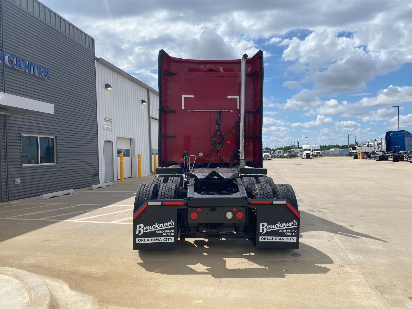 Rear view of a red 2020 Mack AN64T70 sleeper tractor showing the back with mud flaps and chassis. Visible text on mud flaps: 'Bruckner's Oklahoma City' and license plate area. Parts of building and lot in background.