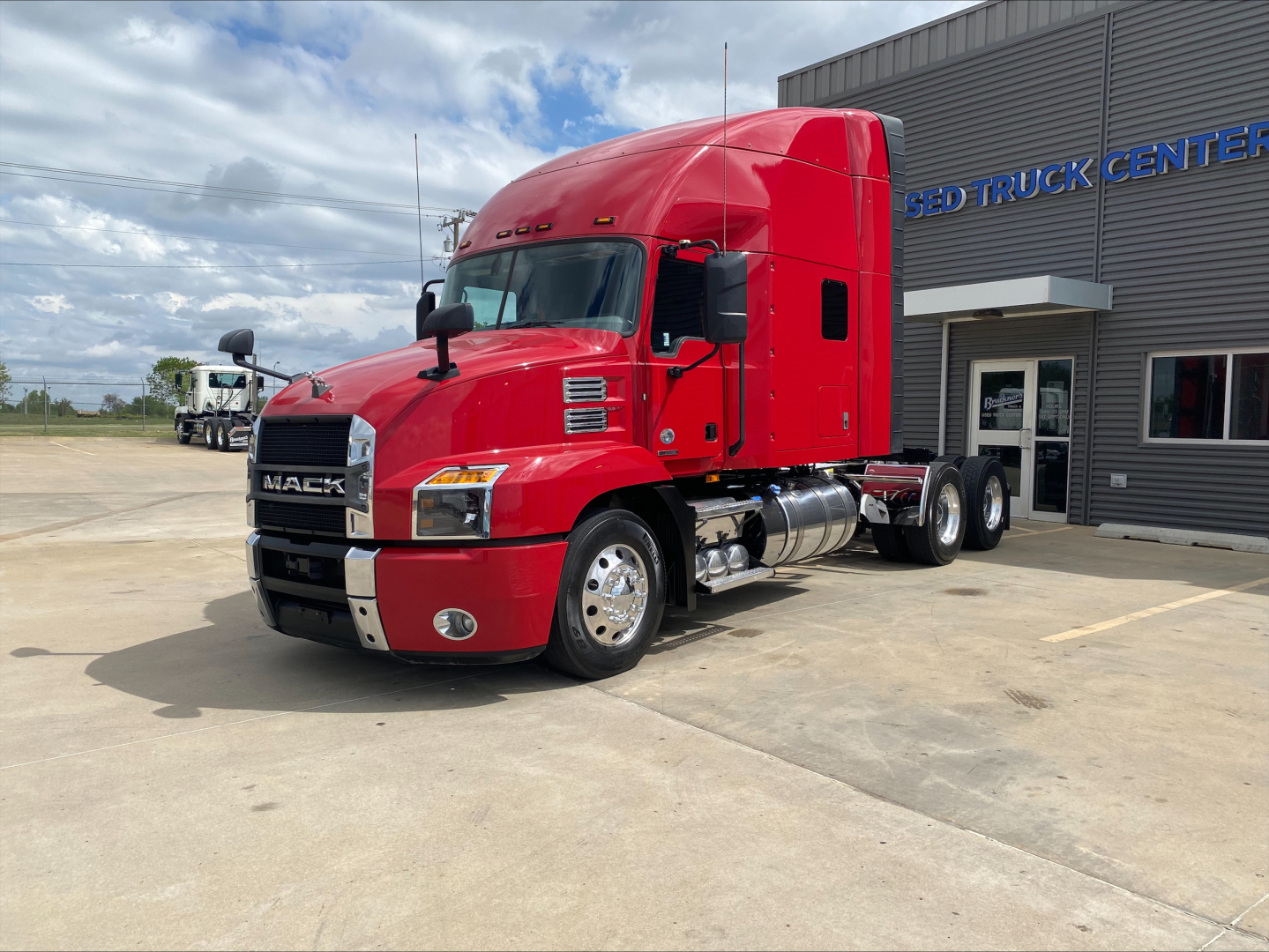 Red 2020 Mack AN64T70 conventional sleeper tractor in front of a dealership, side view with chrome fuel tanks and dual rear axles.