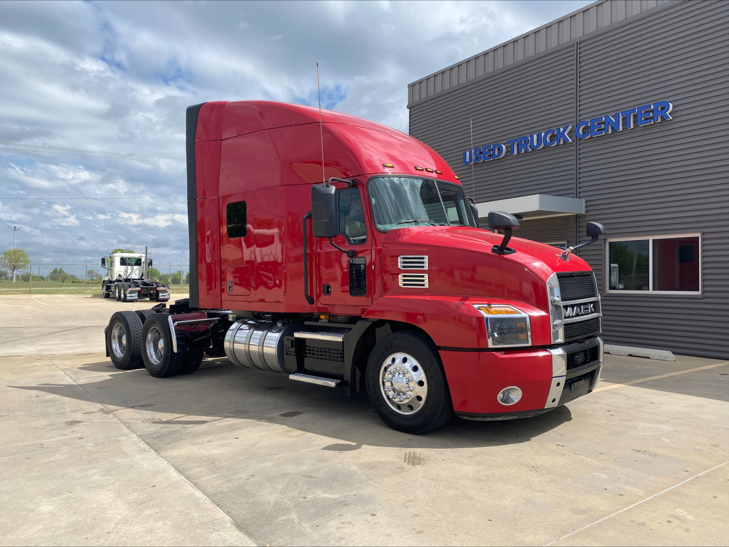 Mack AN64T70 conventional sleeper truck in red parked outside used truck center, side view showing cab with sleeper, dual rear axles, chrome fuel tanks, and large grille.