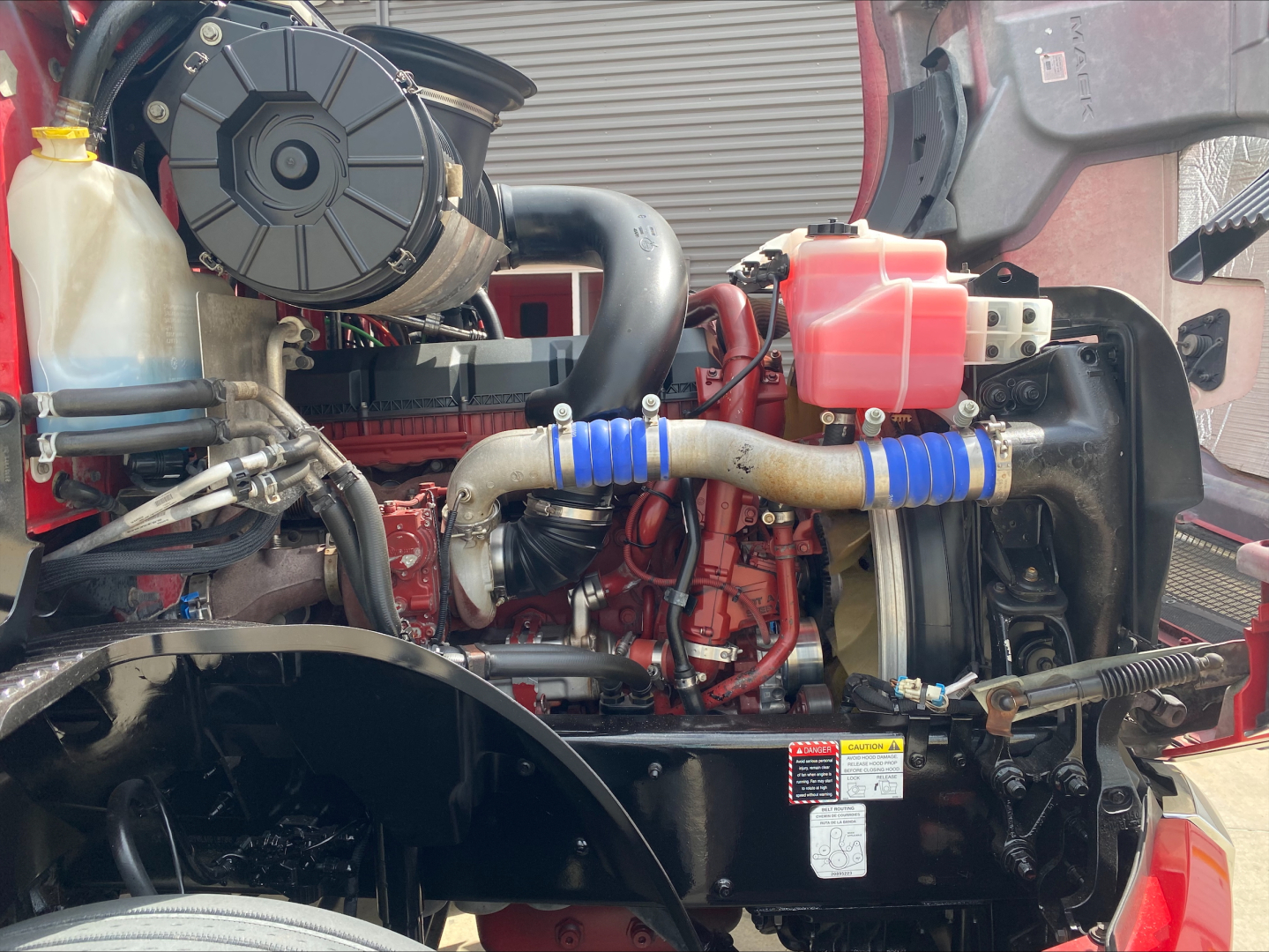 Mack AN64T70 engine bay showing coolant reservoir, red engine block, hoses with blue clamps, air intake, and battery area.