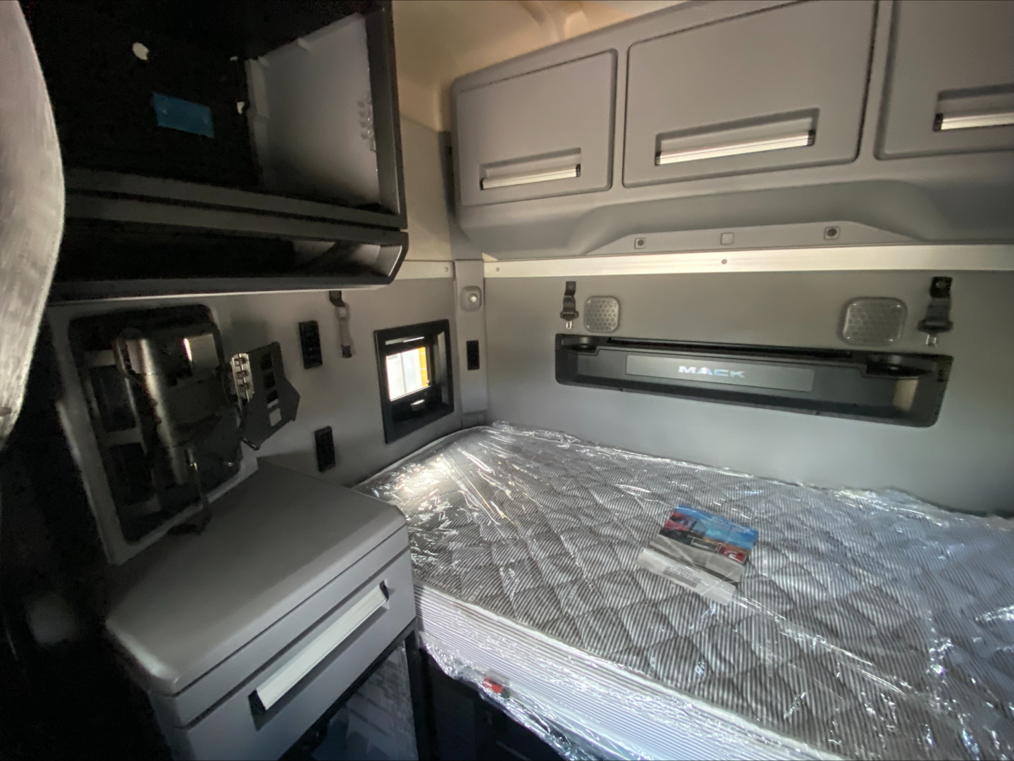Truck cab interior showing sleeper bed and overhead cabinets in a Mack AN64T70, with mattress still wrapped, side panel, and storage compartments.