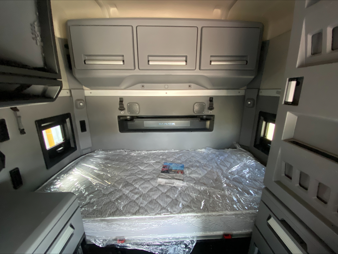 Truck sleeper cab interior with storage cabinets above and around a wrapped mattress in a Mack AN64T70 sleeper, compact dashboard area visible.