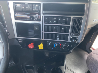 Dashboard of a Mack truck showing gear selector with R N D S M, numerous switches, climate controls, a radio, and two colored knobs (red and yellow) beneath.